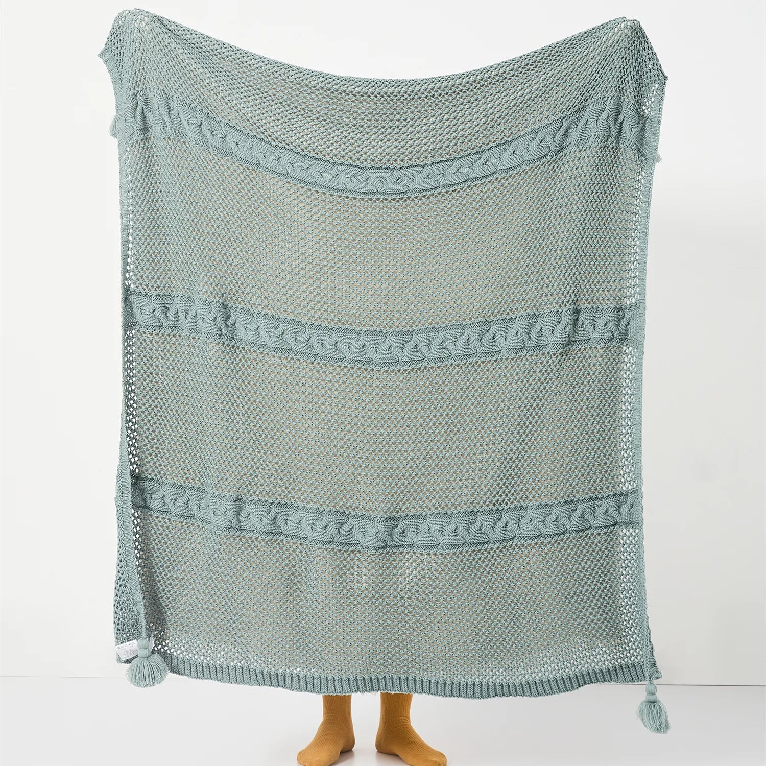 Full view of the large sage-green open-knit throw with horizontal cable stripes and tassels.