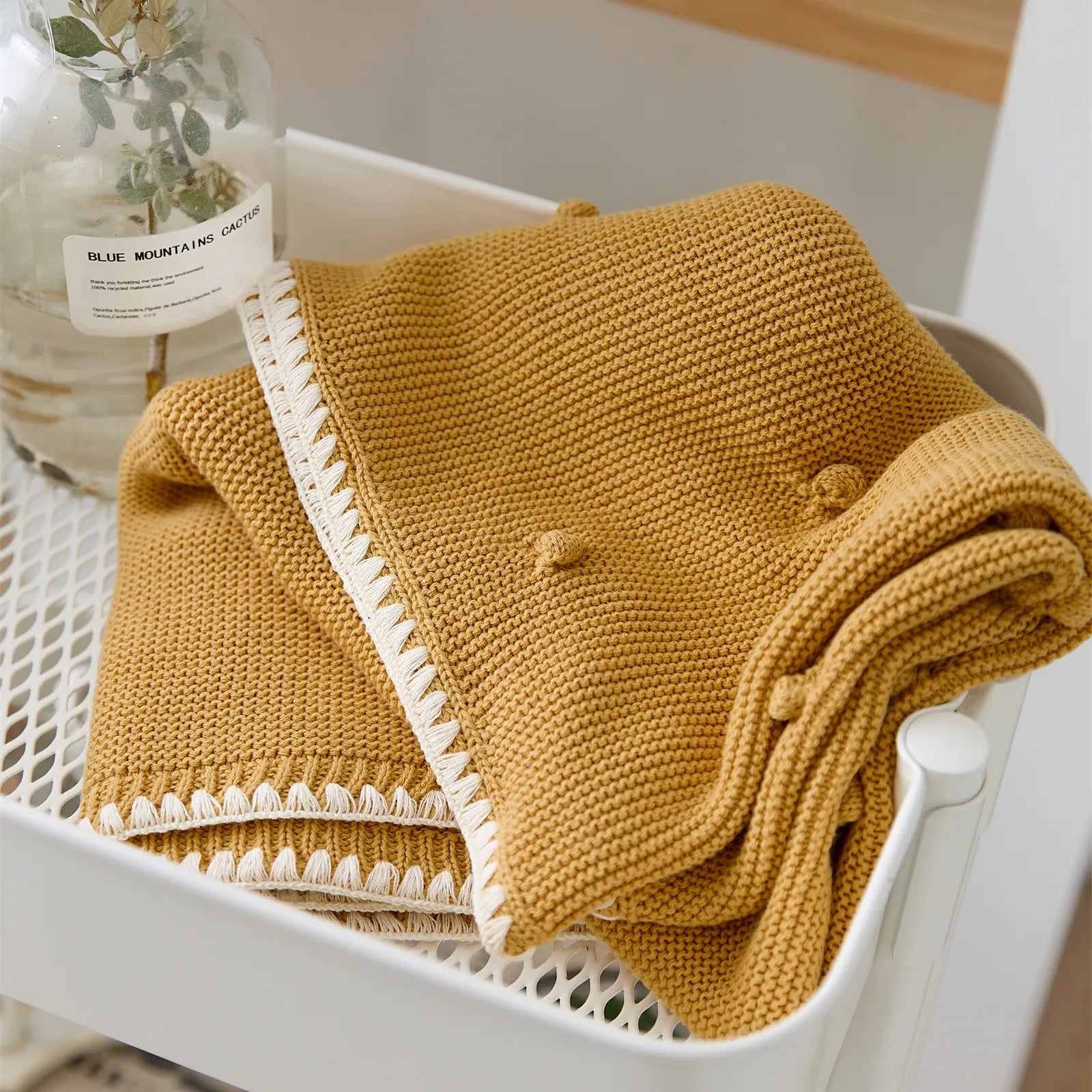 Folded ginger yellow cotton bobble throw placed in a white storage cart next to a glass vase