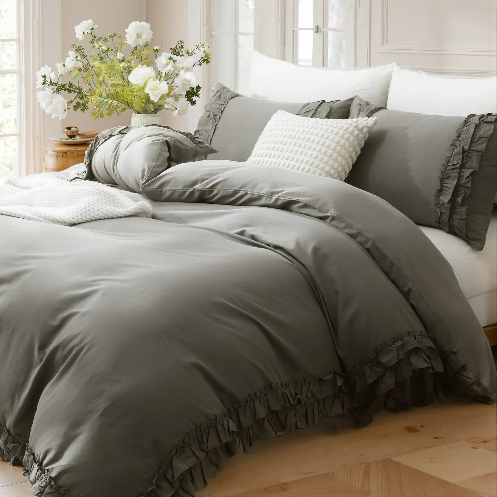 Gray cotton ruffle bedding on a bed with a wooden side table holding a vase of fresh white flowers.