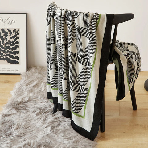 A modern geometric throw blanket in black, white, and subtle green draped over a stylish black chair with a book.