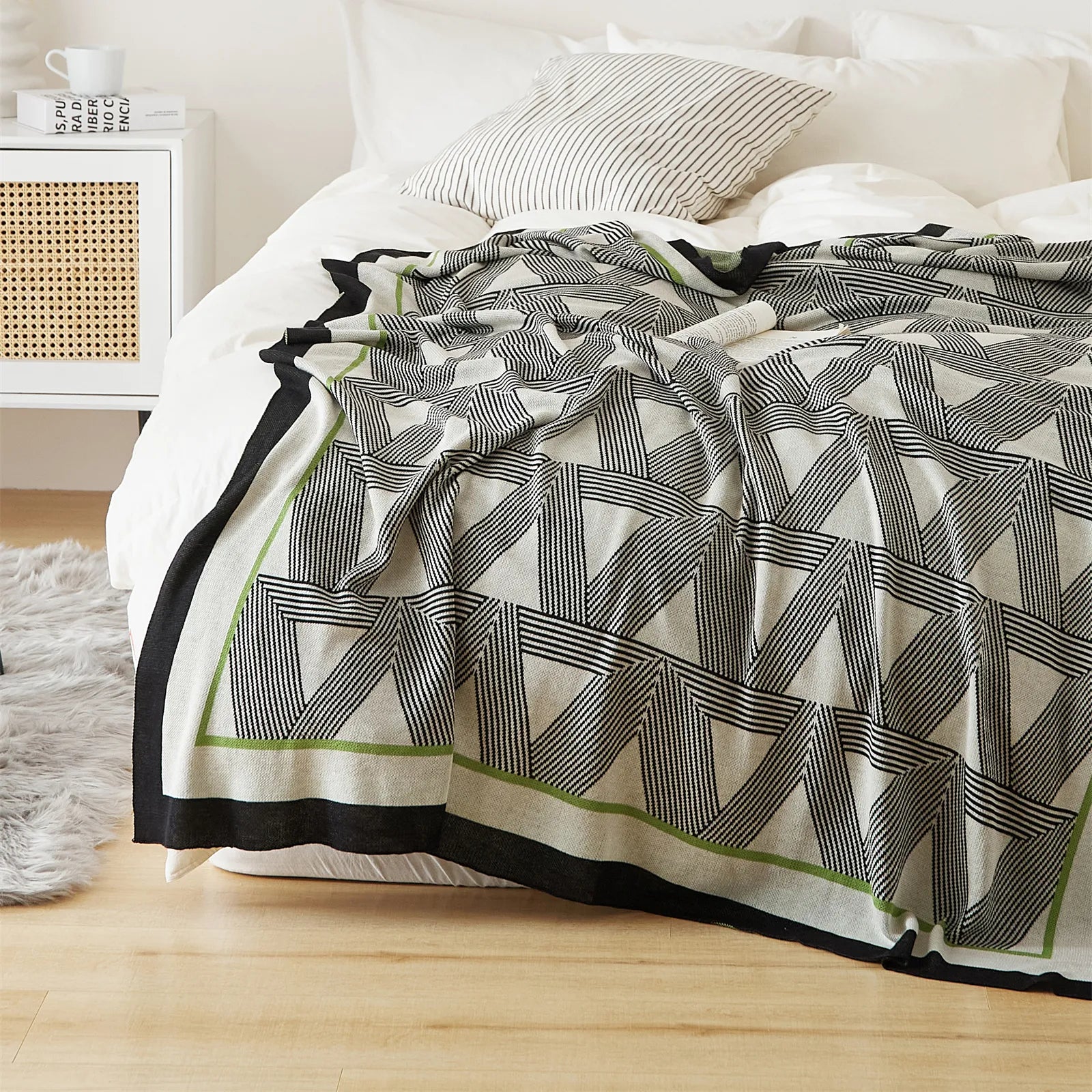 An overhead shot of an elegant geometric throw blanket with green trim, styled on a white bed with a book.