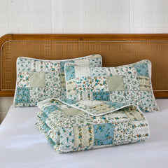 Two pillows and a folded quilt from the green and blue patchwork set on a white bed.
