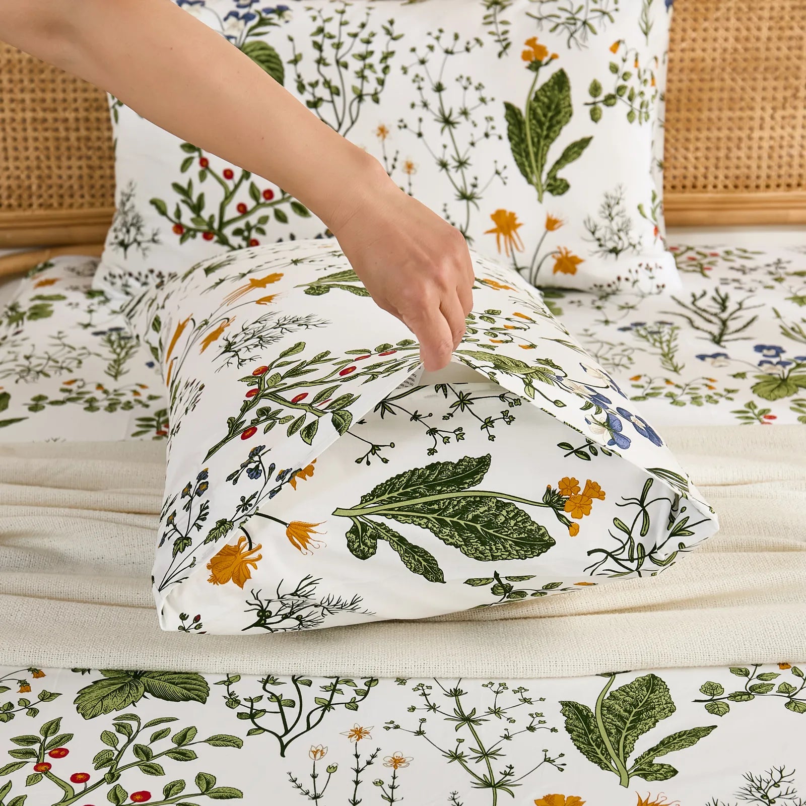 A hand gently pulls open the end of a white cotton botanical print pillowcase, revealing the inner details and quality.
