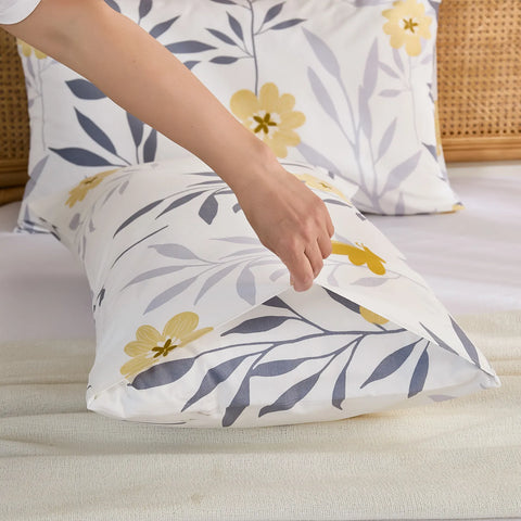A hand adjusts a white cotton pillowcase with a yellow floral and grey leaf print, showing the product's envelope closure.