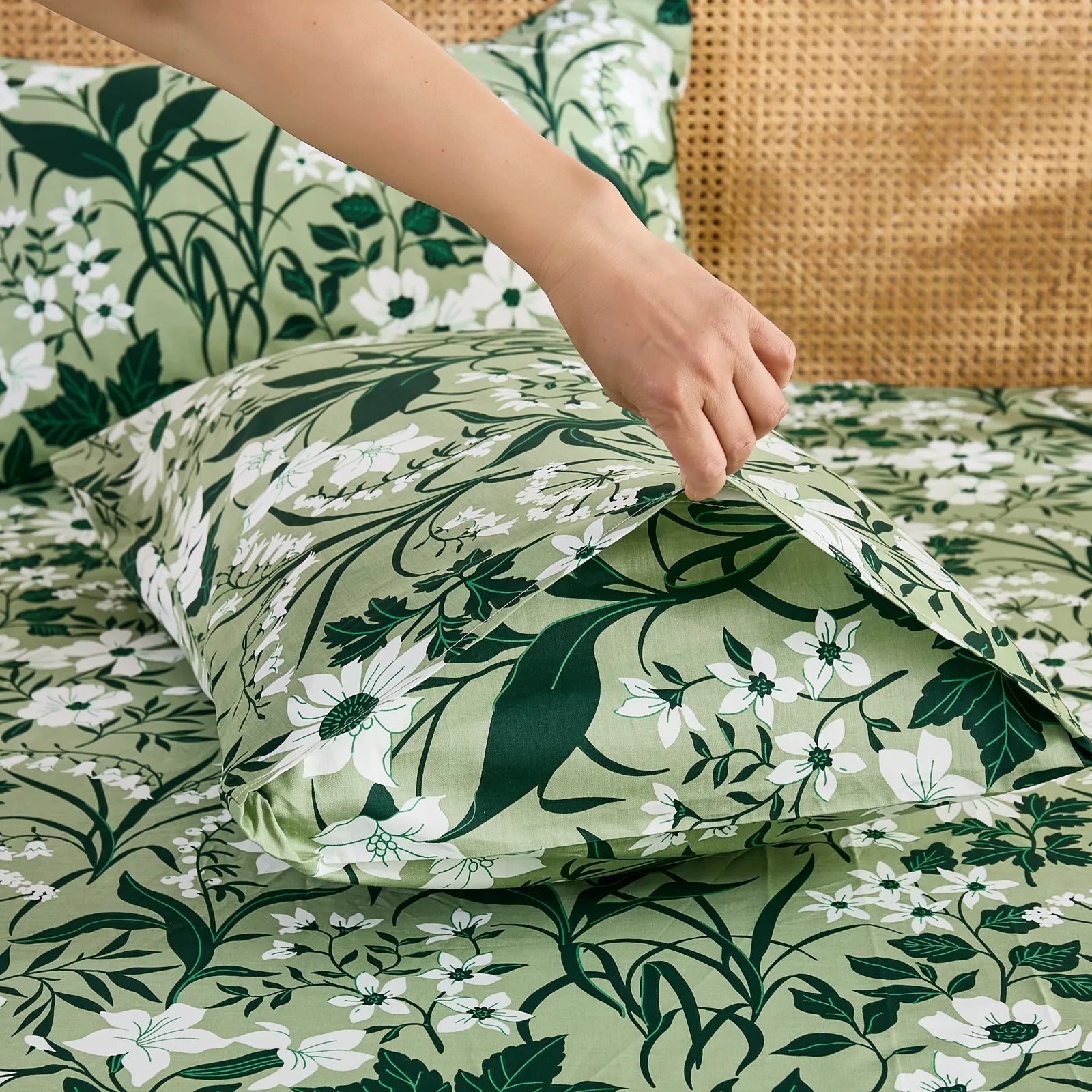 A hand gently pulls open the end of an olive green cotton pillowcase, showing the deep green floral and white flower pattern and envelope closure.