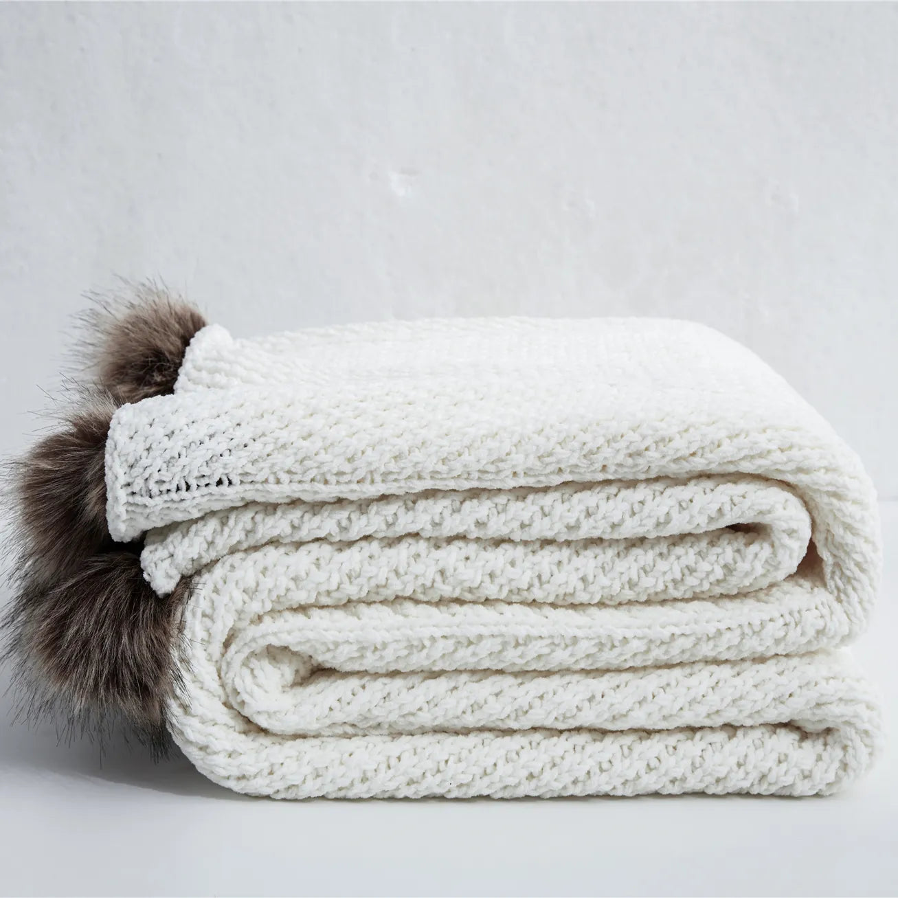 Folded ivory chunky knit throw blanket with brown faux fur pom-poms
