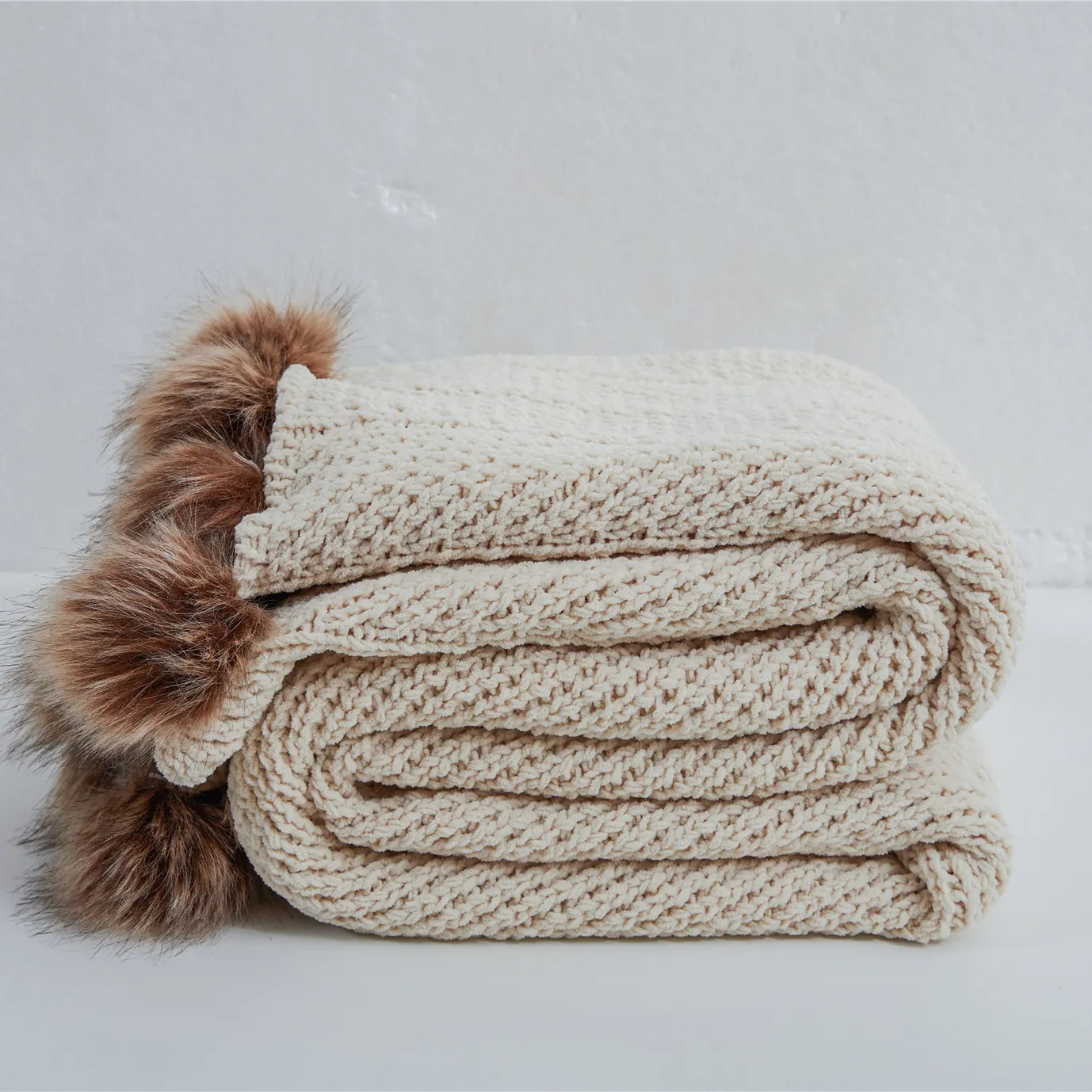 Folded light beige chunky knit throw blanket with brown faux fur pom-poms
