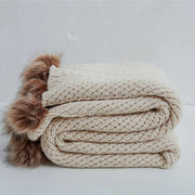 Folded light beige chunky knit throw blanket with brown faux fur pom-poms