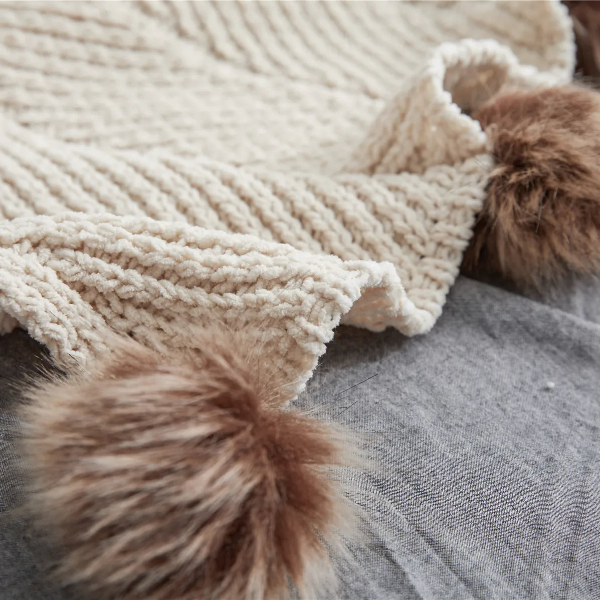 Detail of light beige cotton knit fabric corner with large brown faux fur pom-pom on grey background
