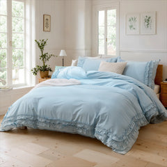 Light blue cotton double ruffle duvet cover and pillows on a wooden bed with a nightstand and lamp.