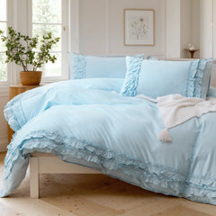 Light blue cotton duvet cover with pillows and a knit throw blanket, beside a small table with a plant.