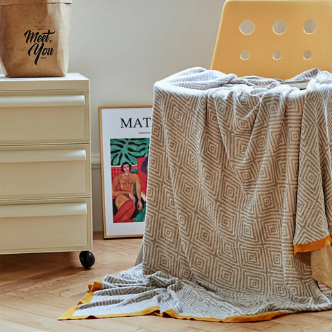 Light-grey diamond pattern throw with a mustard border draped over a modern yellow chair in a bright indoor corner.