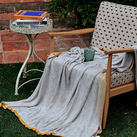 Light-grey diamond pattern throw with a mustard border draped over a wooden armchair in an outdoor garden setting.