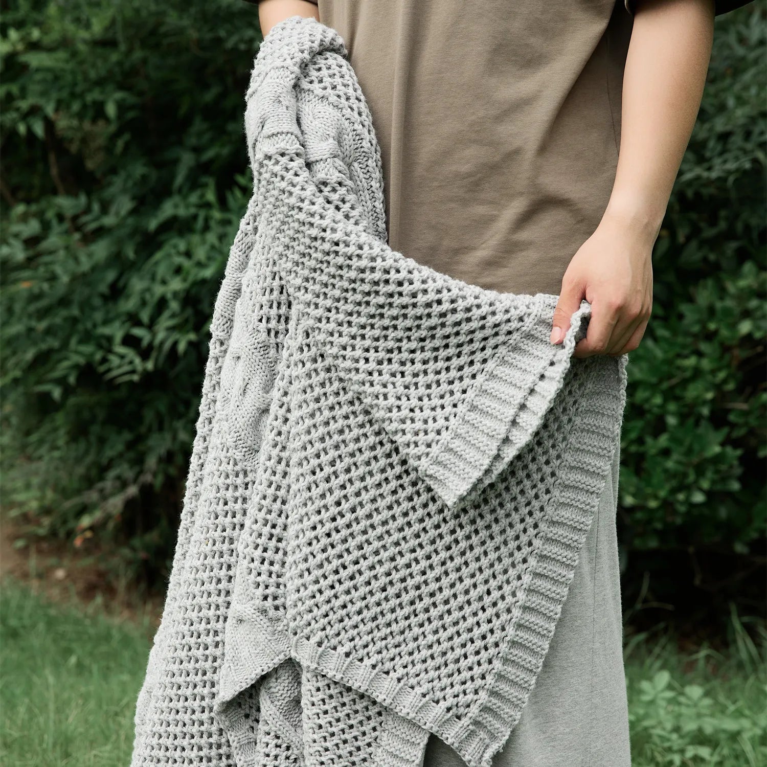 Person holding a light-grey knit throw blanket outdoors, highlighting the mesh texture and color.