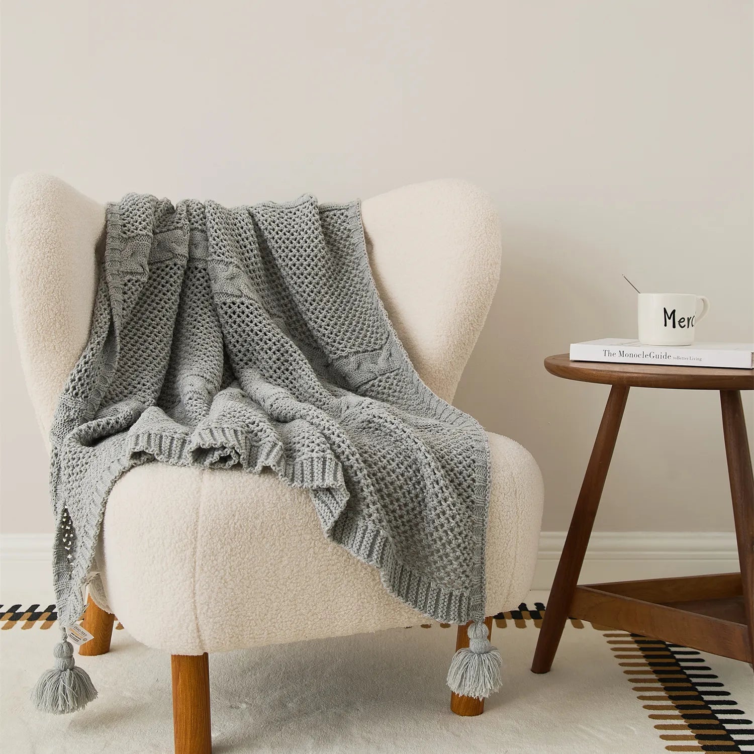 Light-grey knit throw with tassels draped over a cream teddy armchair next to a wooden side table.