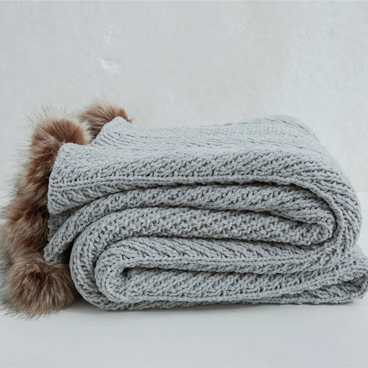 Folded light heather grey chunky knit throw blanket with brown faux fur pom-poms