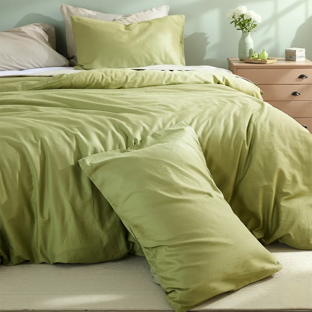 Matcha green duvet with casually stacked pillows beside a pink nightstand holding a vase of white flowers and green fruit.