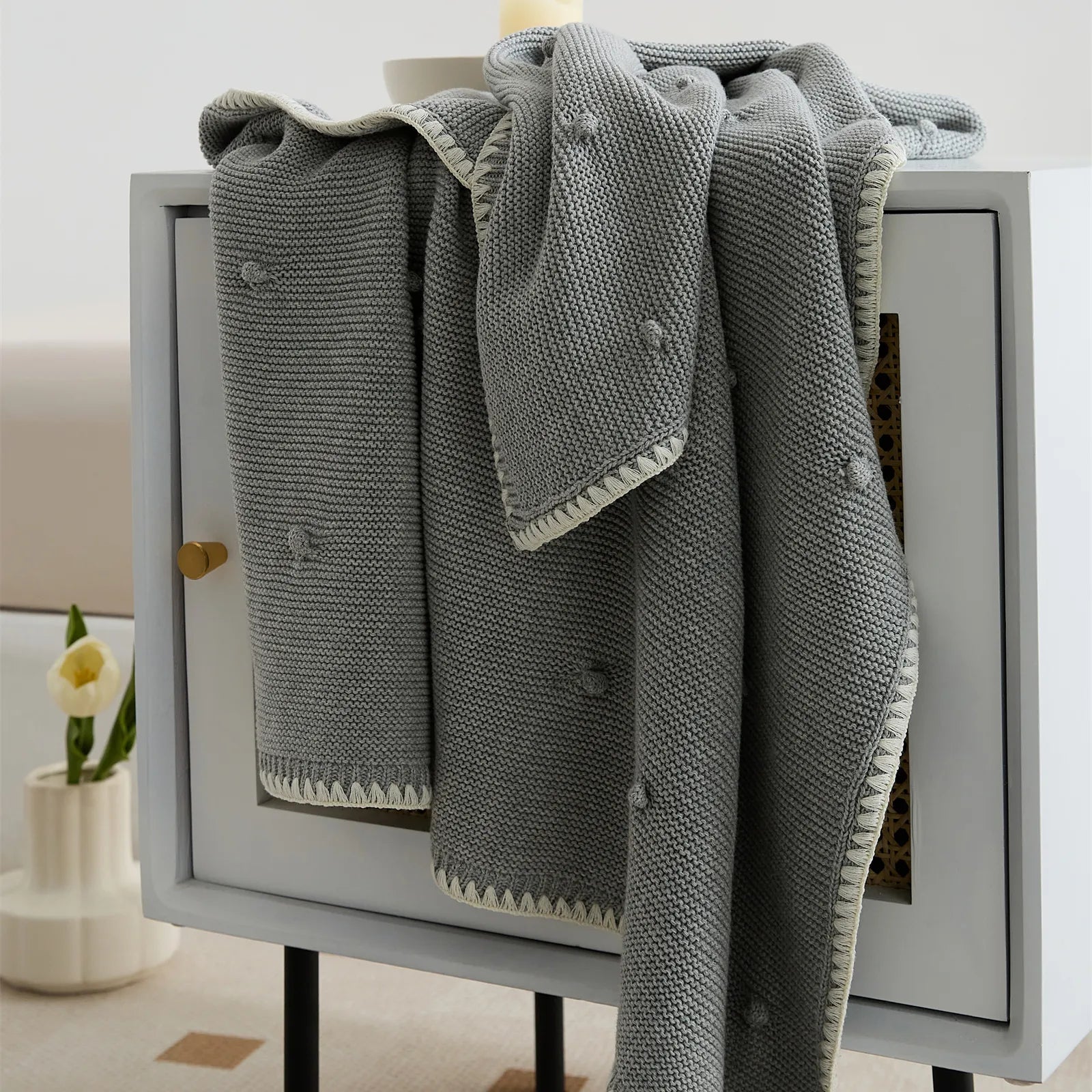 Medium grey cotton bobble throw draped over a modern white and rattan bedside cabinet