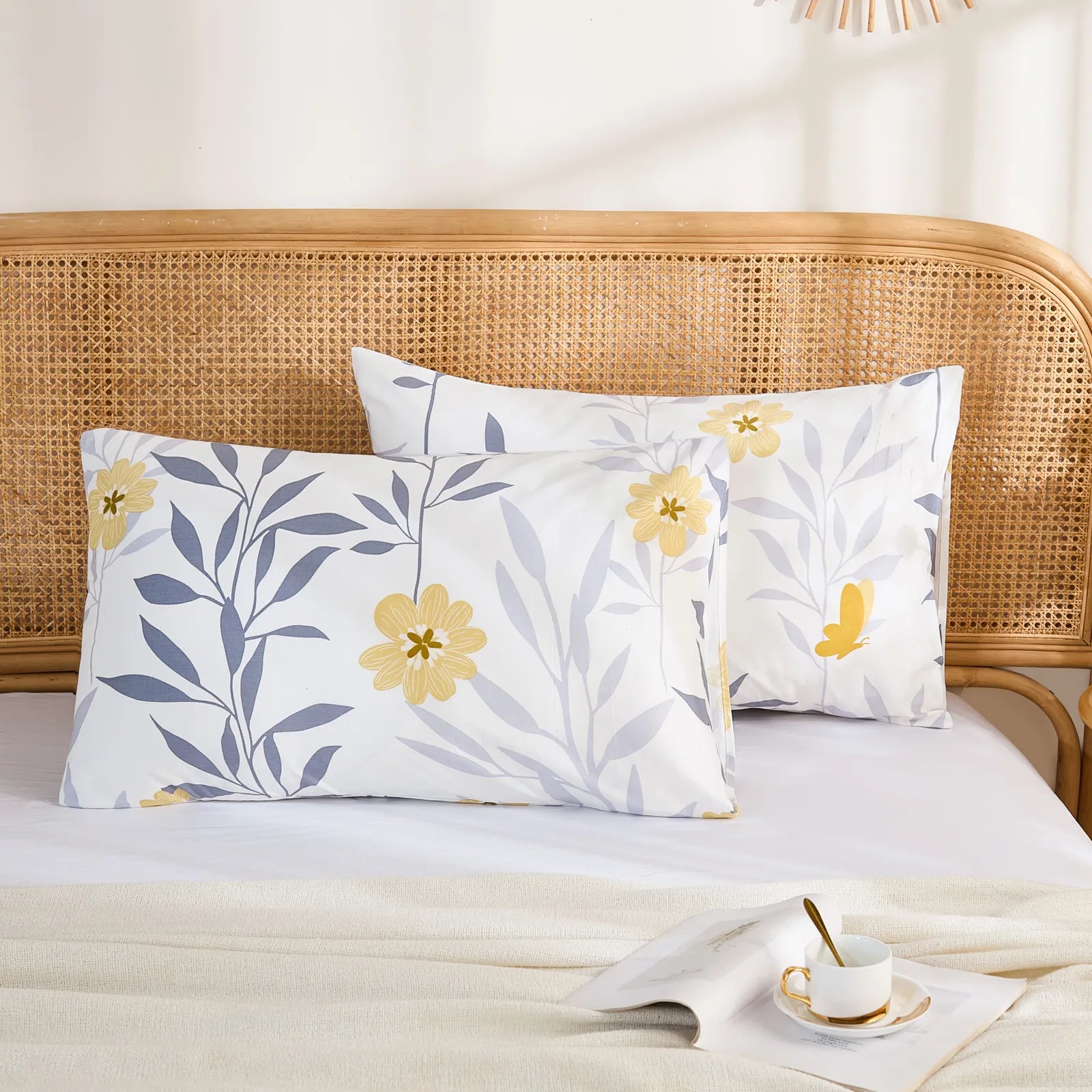 A pair of modern yellow floral and grey leaf print cotton pillowcases, resting on a clean white bed with a woven headboard.