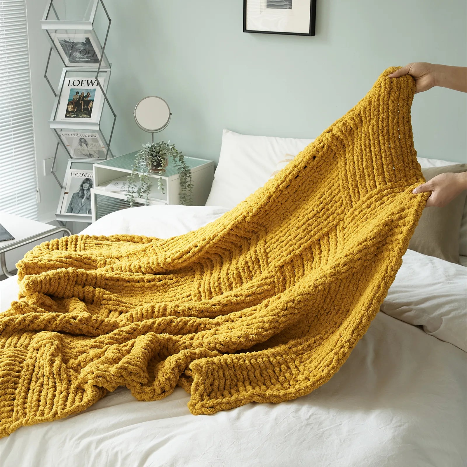 A hand holds up a mustard yellow chunky knit blanket, showcasing its vibrant color and thick texture on a bed.