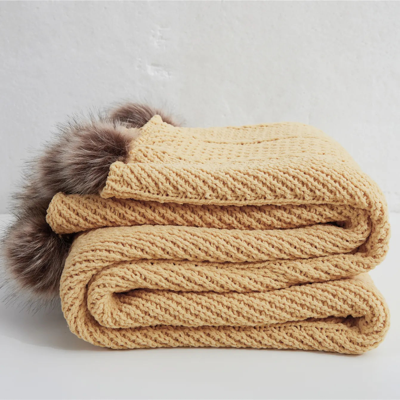 Folded mustard yellow cotton chunky knit throw blanket with brown faux fur pom-poms