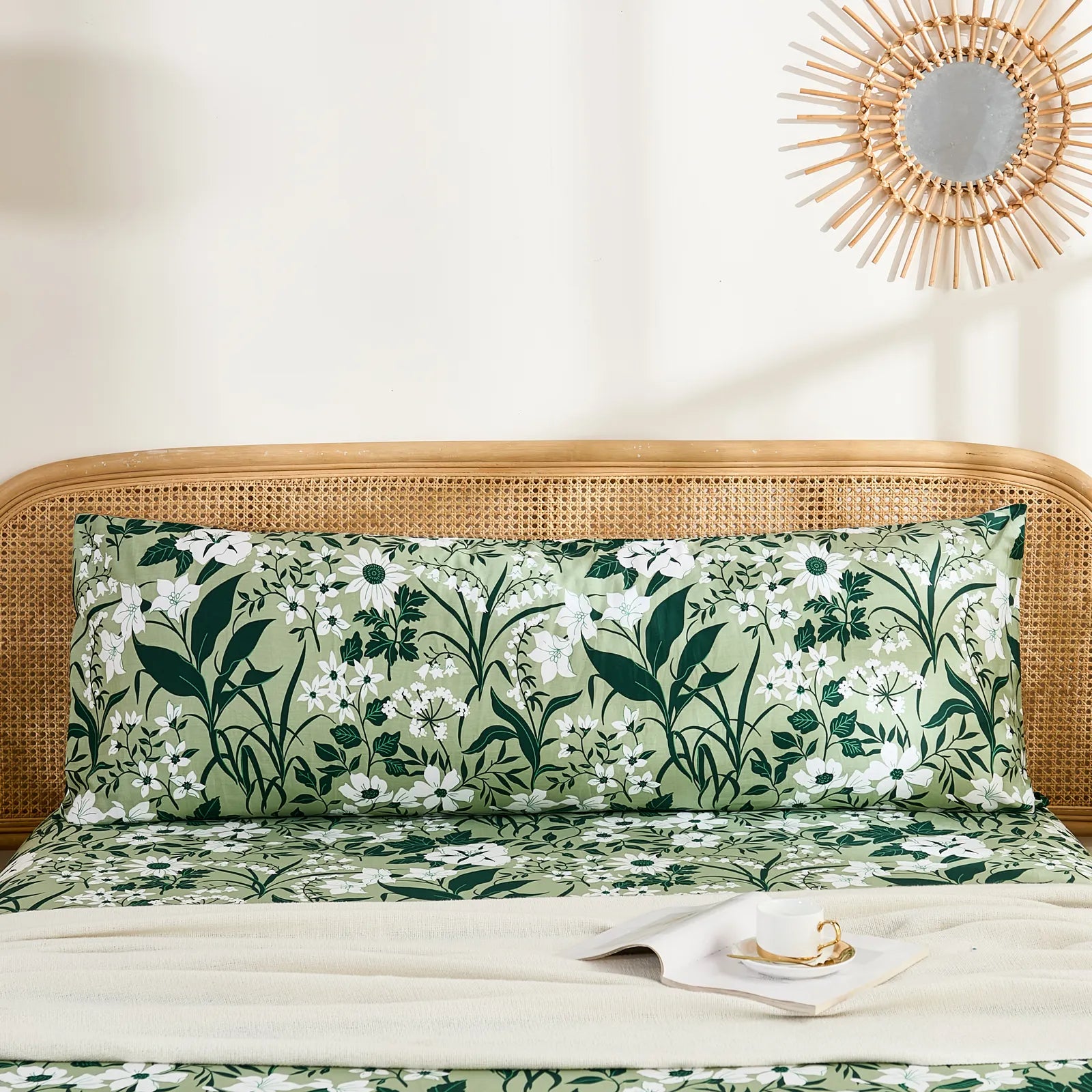A single long cotton body pillow in an olive green and white floral print, positioned against a rattan headboard in a cozy bedroom setting.