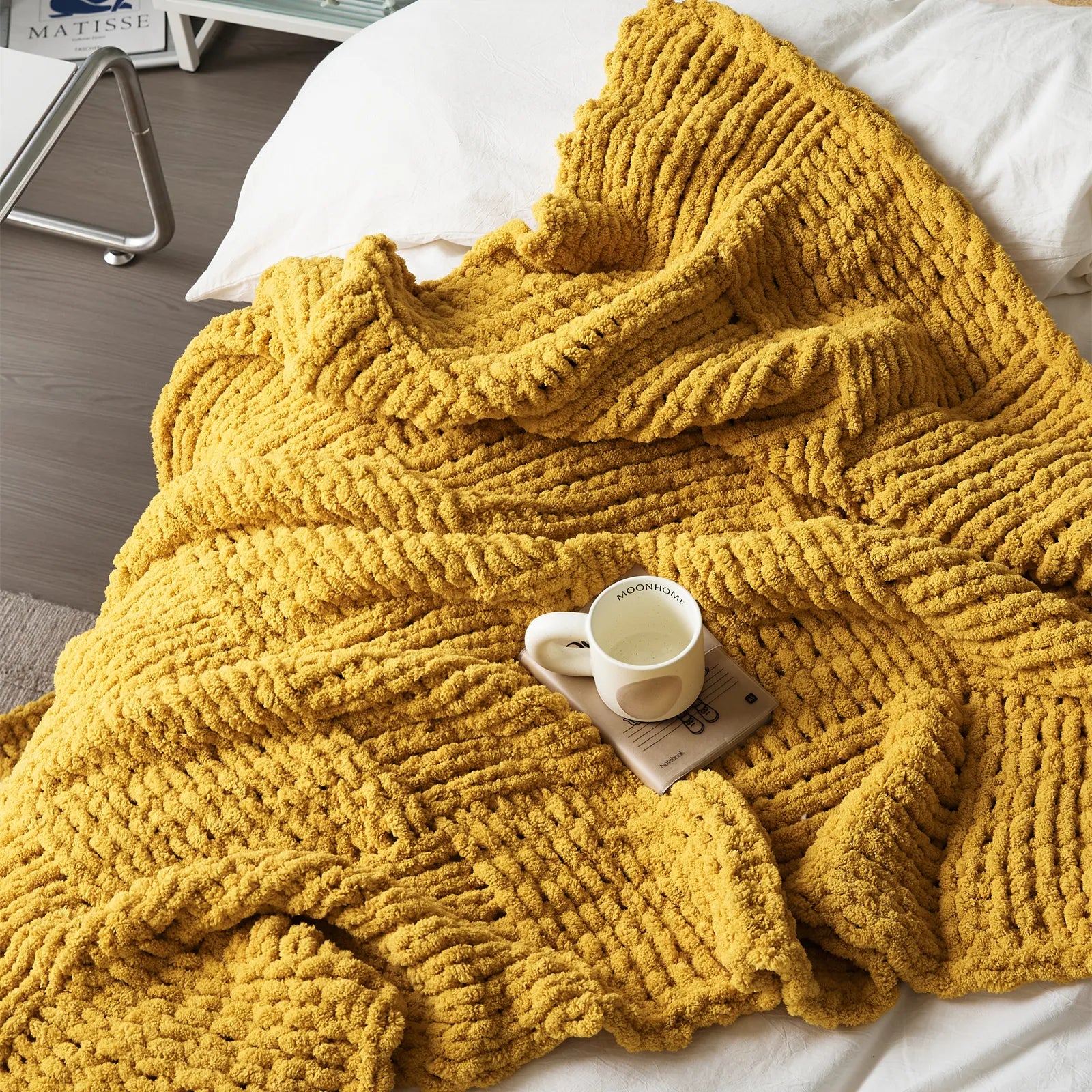 An overhead view of a soft mustard yellow chunky knit blanket lying on a bed with a mug and book, highlighting its inviting texture.
