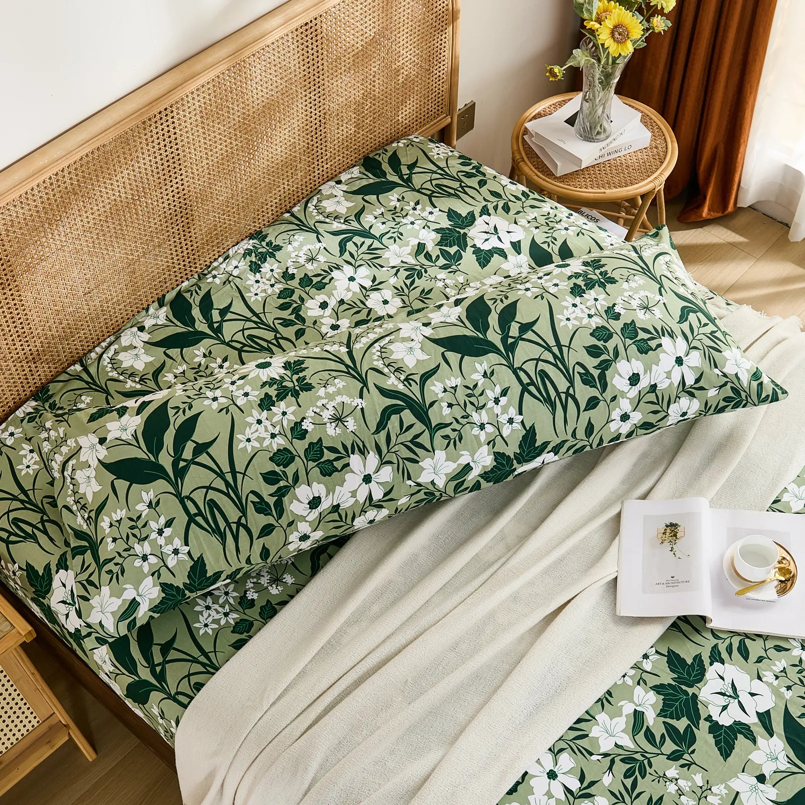 An overhead view of a long cotton body pillow in olive green floral print, laid across a bed with matching sheets and a white throw.