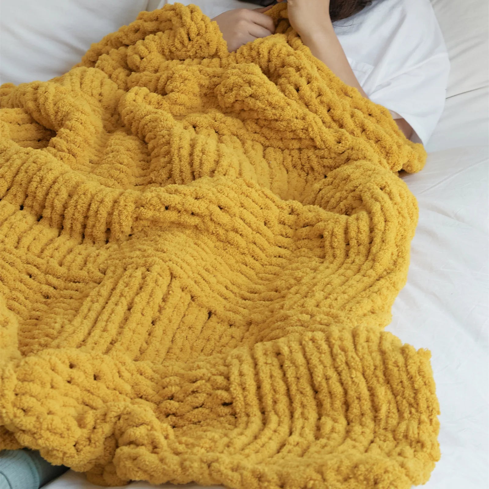 A person is wrapped in a mustard yellow chunky knit blanket on a bed, highlighting its warmth and comfort.