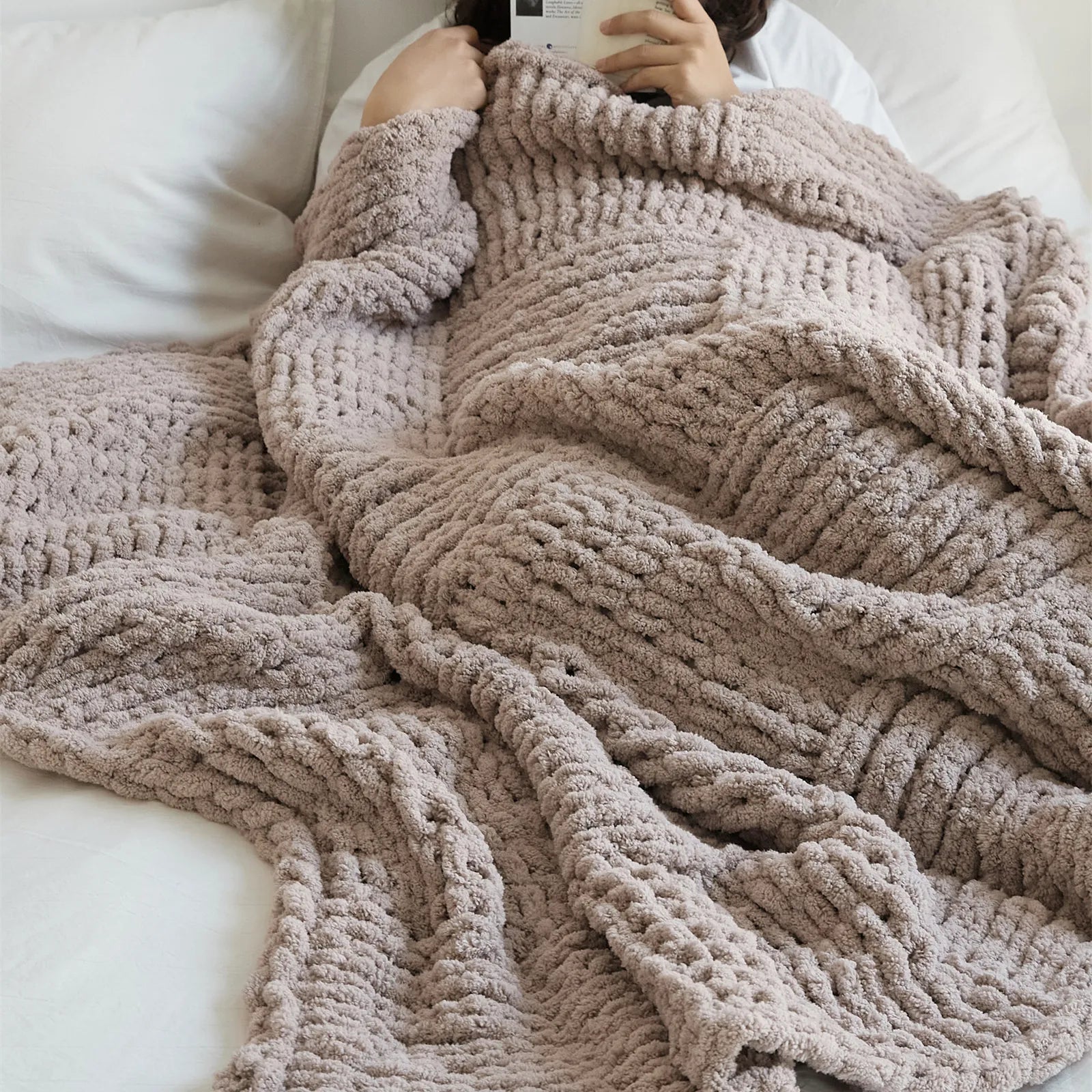 A person is comfortably wrapped in a soft taupe chunky knit blanket, highlighting its warmth and luxurious feel