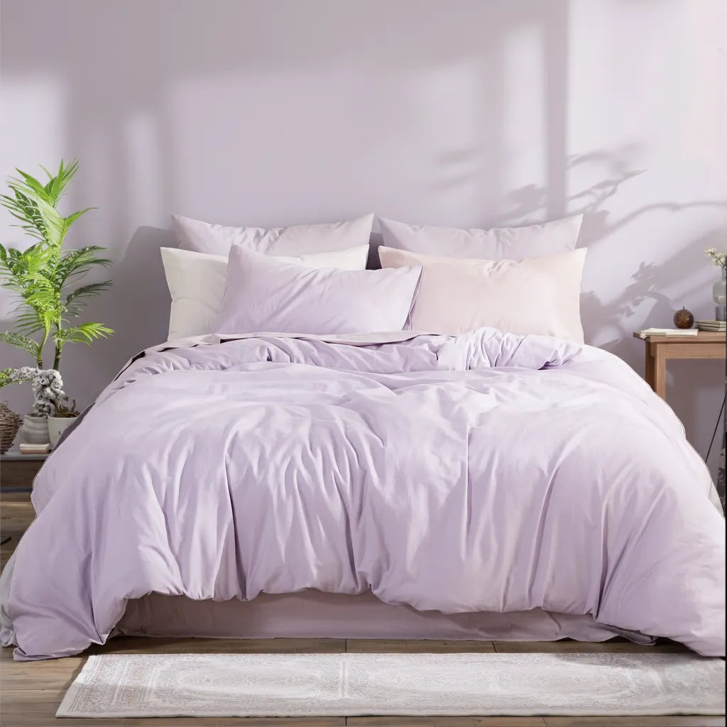 Purple solid cotton duvet with four pillows in soft sunlight casting tree shadows, complemented by a dark wood floor and areca palm.