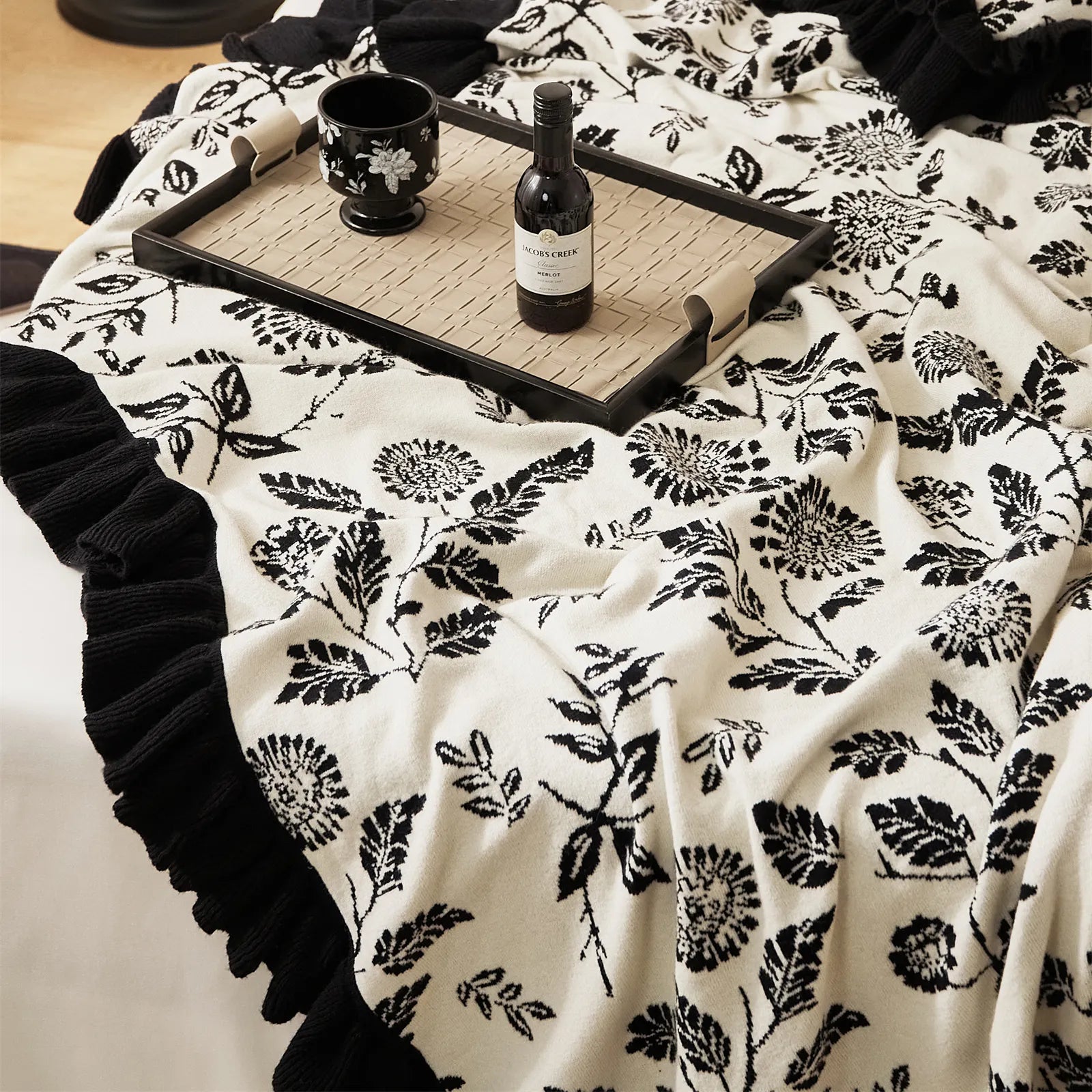 An elegant black leaf patterned microfiber blanket with a ruffled edge, spread out on a crisp white bed.