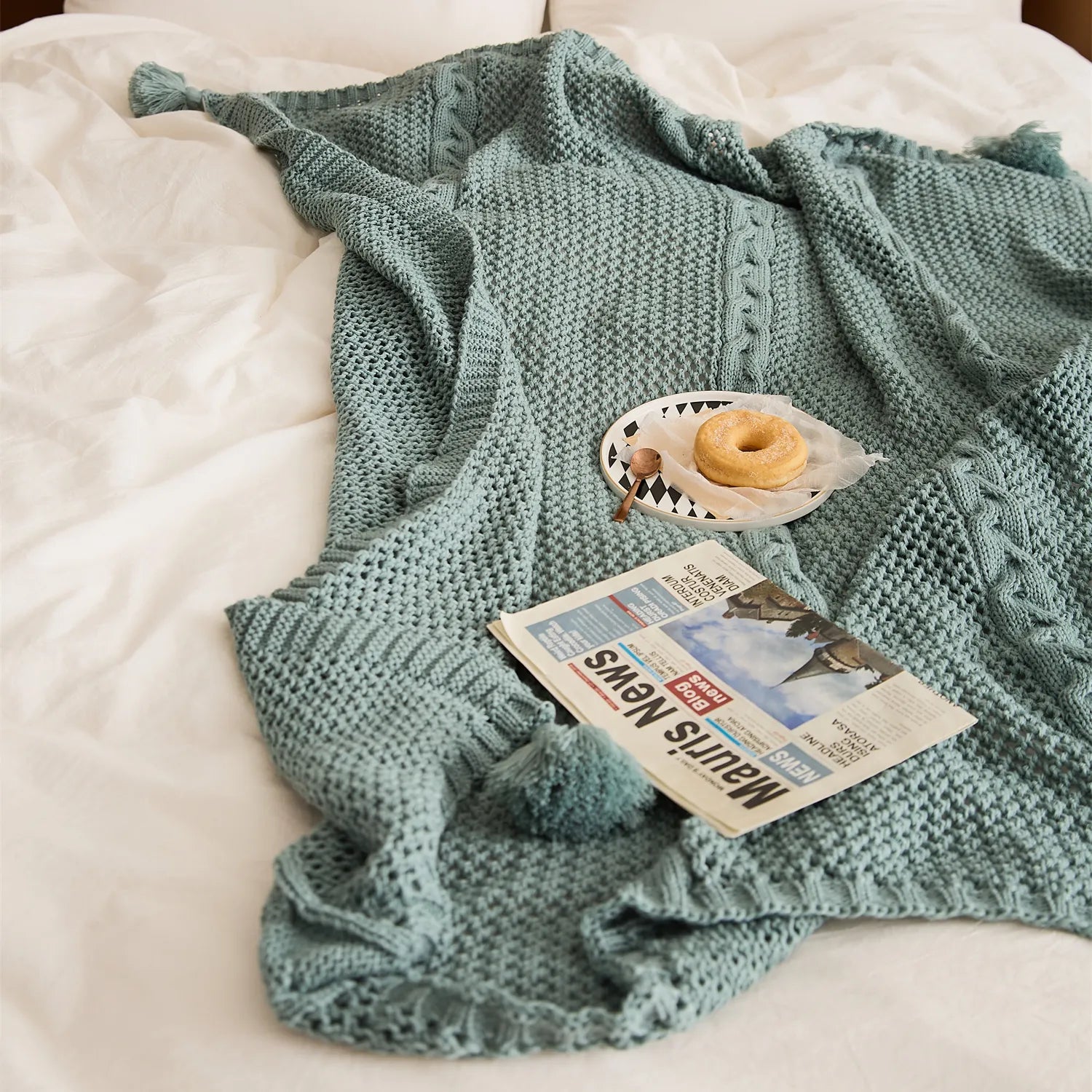 Close-up of sage-green knit throw on a white bed with a donut and newspaper, featuring cable and open-knit textures.