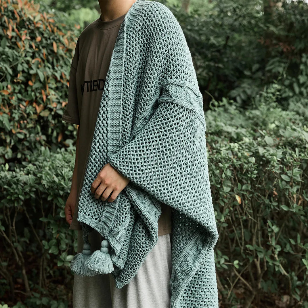 Person wrapped in a sage-green knit throw blanket with tassels, showcasing its size and drape in an outdoor setting.