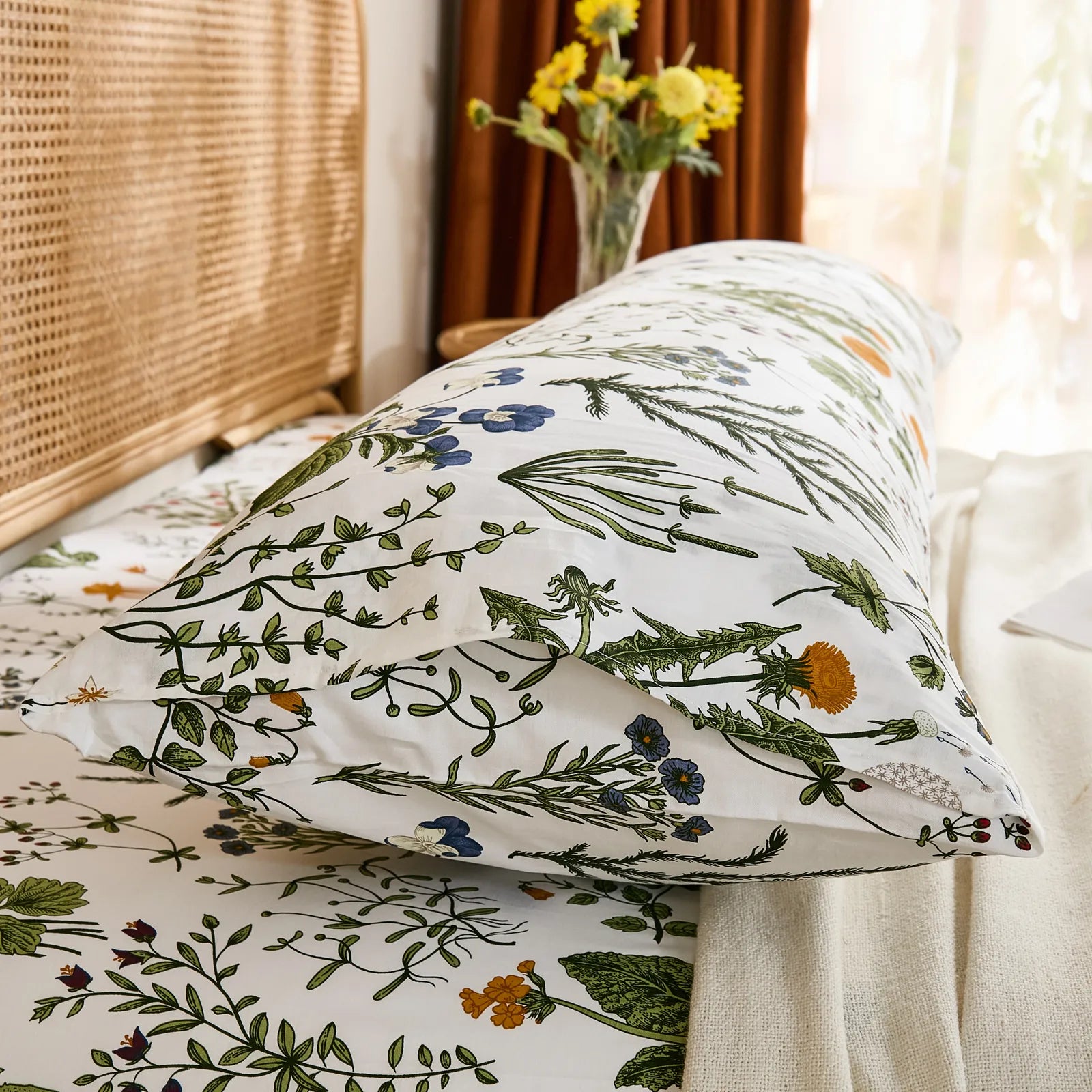 A side view of a white cotton body pillow with a botanical print, propped up on a bed near a window with sheer curtains.