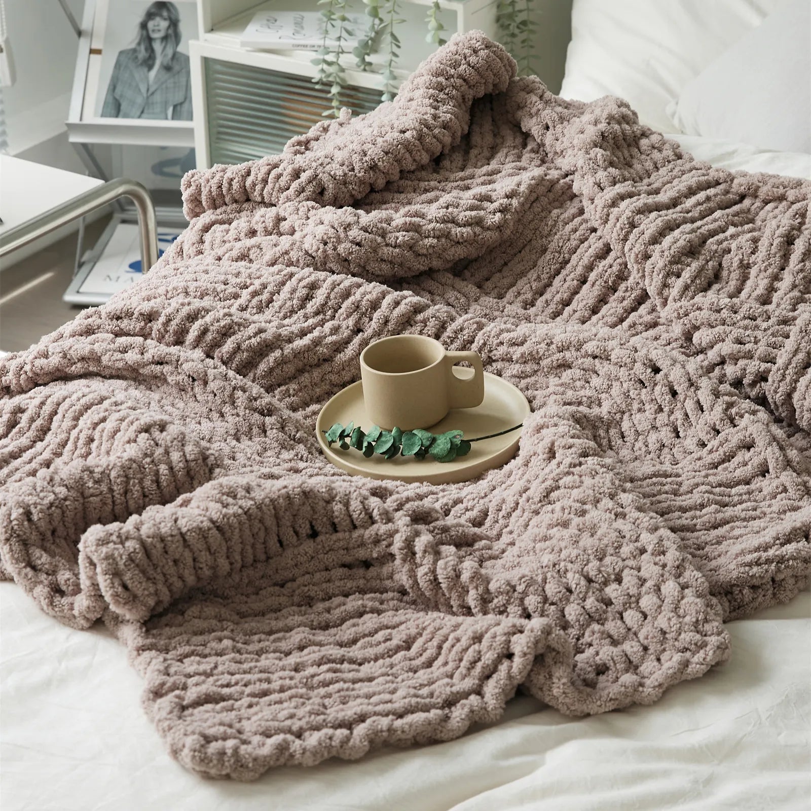 A plush taupe chunky knit blanket is spread out on a white bed, with a mug on a tray, creating a warm, cozy scene.