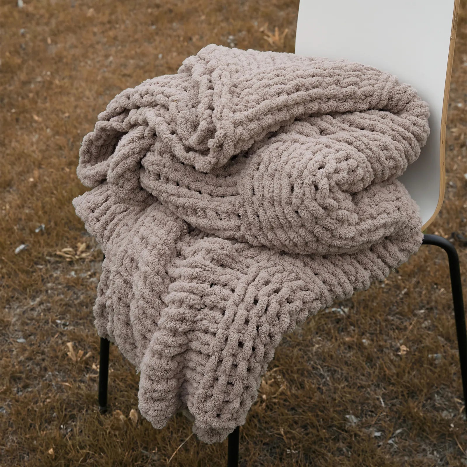A cozy taupe chunky knit blanket is neatly folded on a chair outdoors, showing its soft, thick texture.