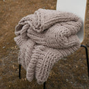 A cozy taupe chunky knit blanket is neatly folded on a chair outdoors, showing its soft, thick texture.