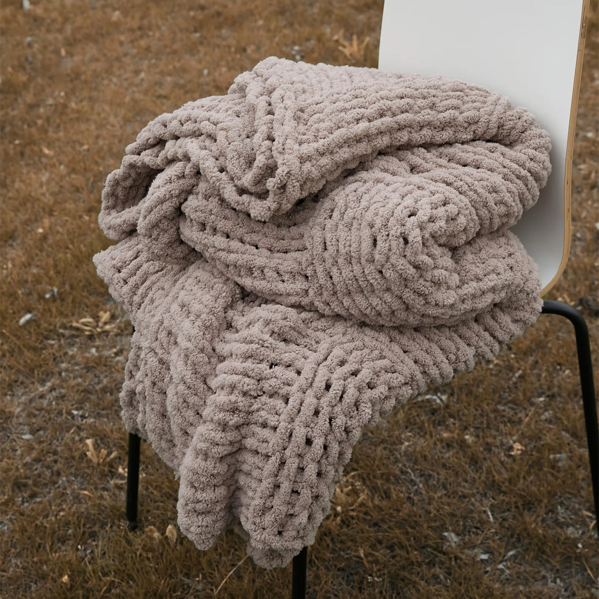 A cozy taupe chunky knit blanket is neatly folded on a chair outdoors, showing its soft, thick texture.