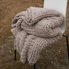 A cozy taupe chunky knit blanket is neatly folded on a chair outdoors, showing its soft, thick texture.