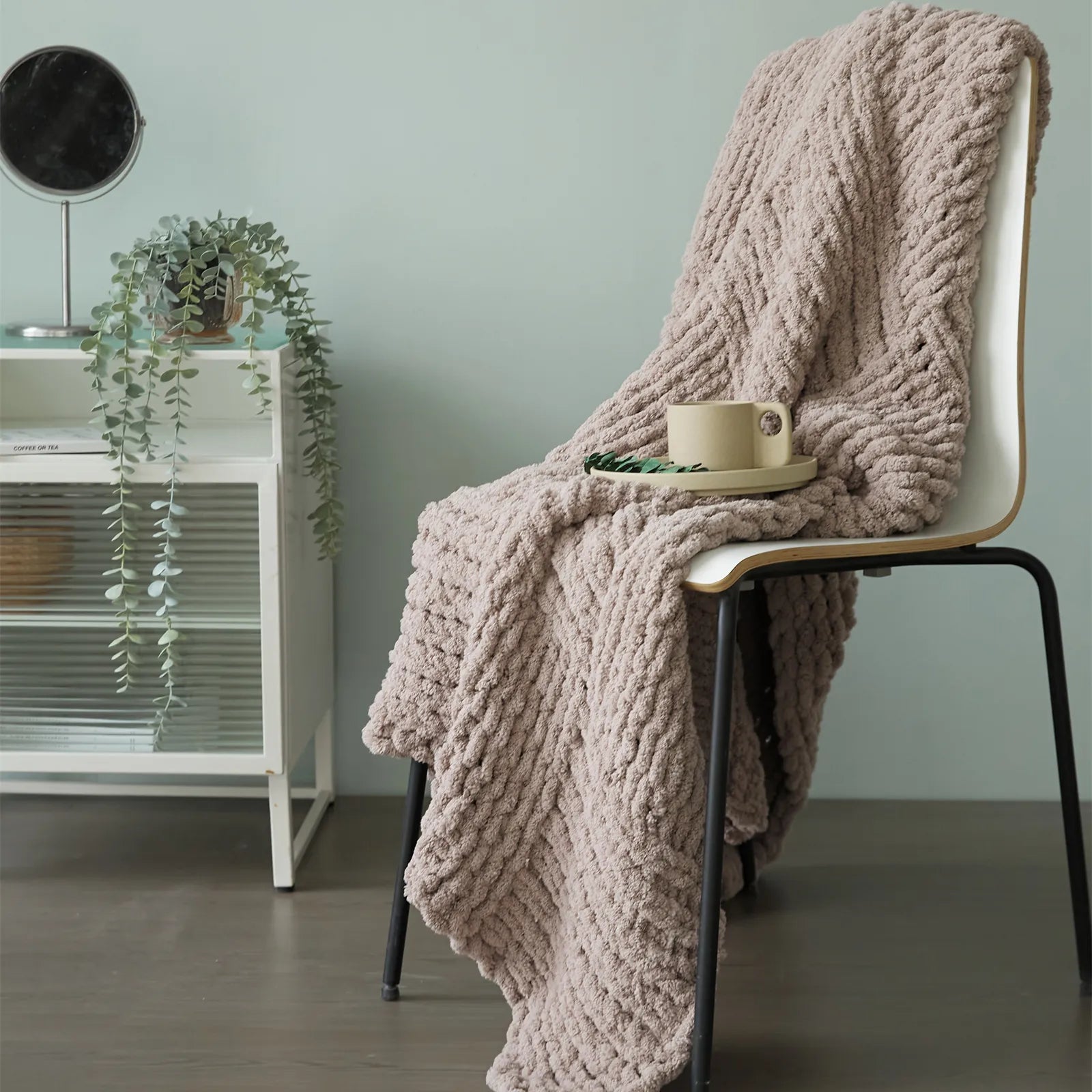 A chunky taupe knit blanket is draped over a chair, styled with a coffee cup and plant for a modern, inviting look.