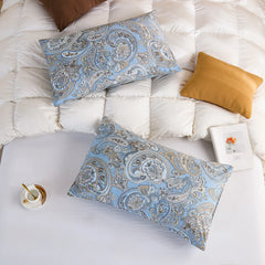 An overhead shot of a bedding set featuring two light blue paisley print cotton pillowcases on a white quilted comforter, with accent pillows.