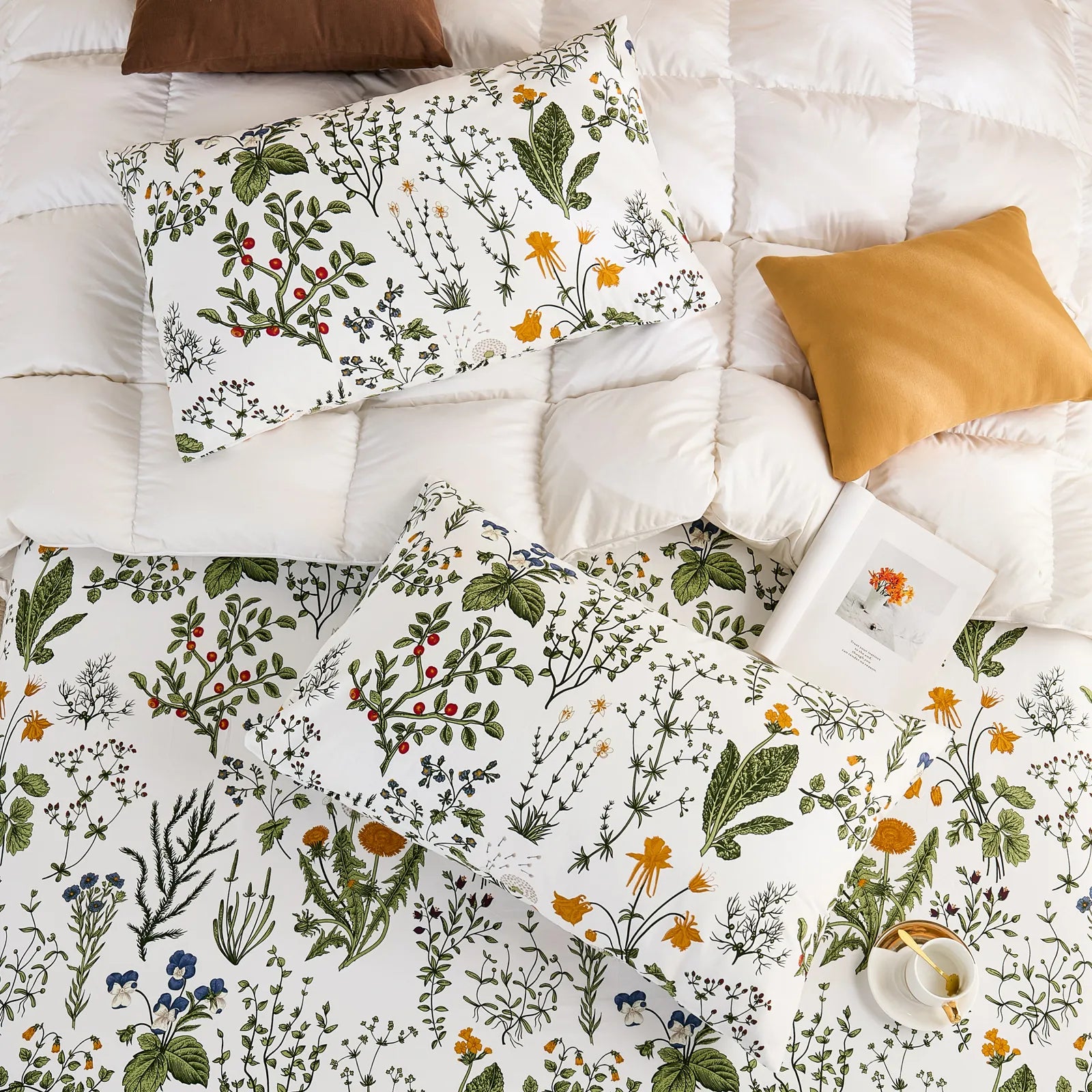 An overhead shot of a bedding set including a white cotton botanical pillowcase, a matching sheet, and a fluffy white comforter, with accent pillows.