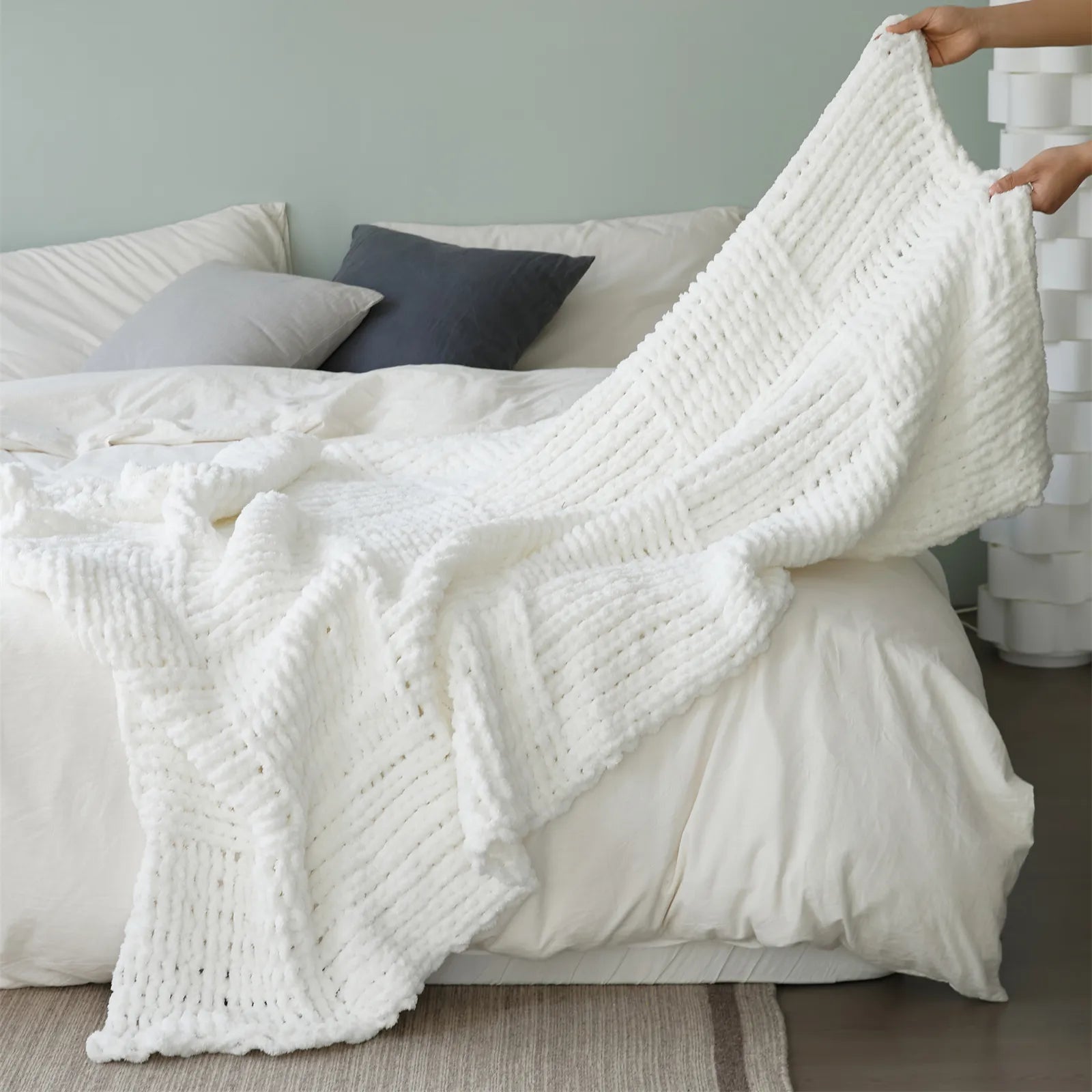 A hand gently spreading a cozy, chunky knit throw blanket in a clean white color across a bed, highlighting its texture and softness.